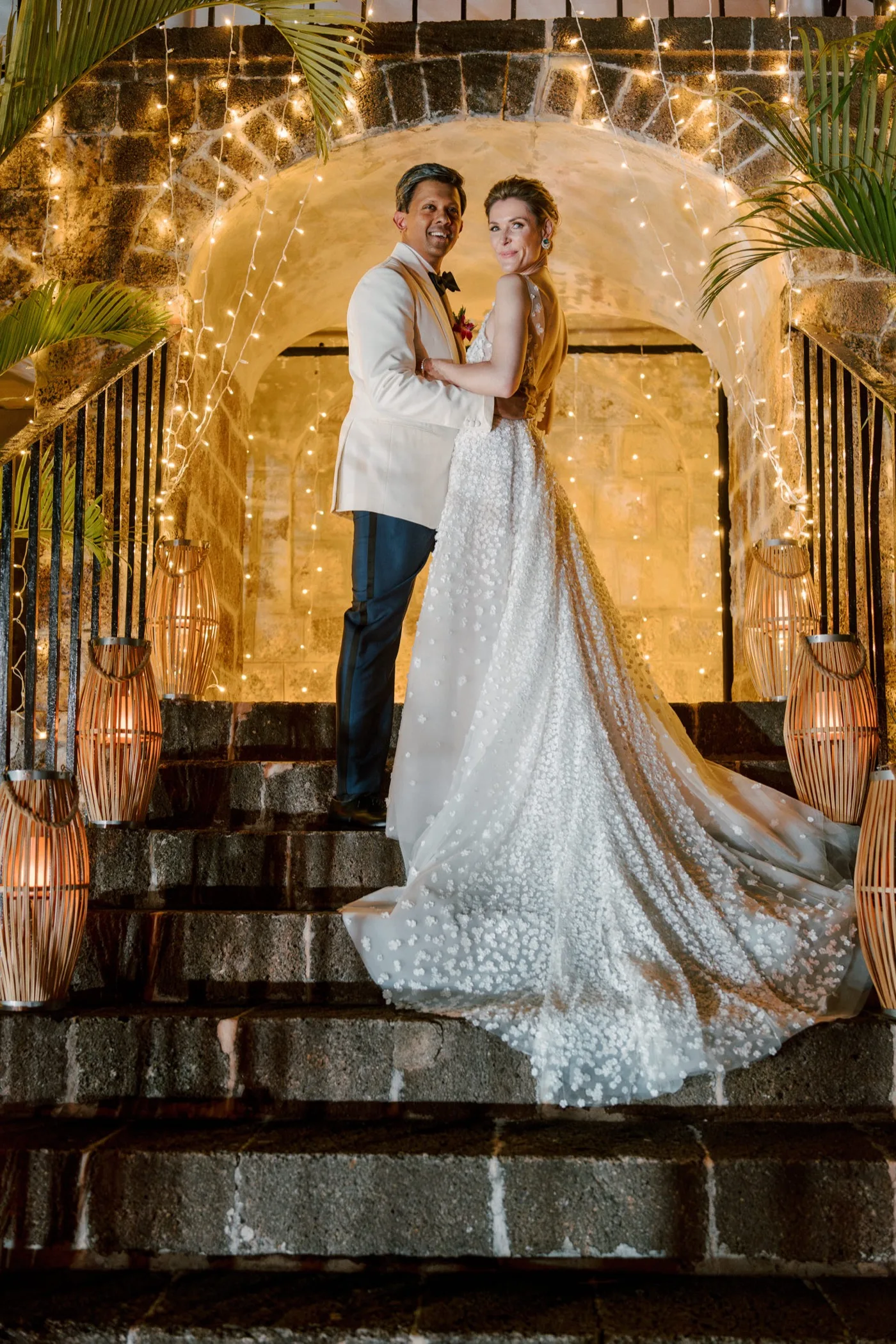 Couple embracing on fairy-lit stone steps beneath an arched entrance — wedding photography by David Josué