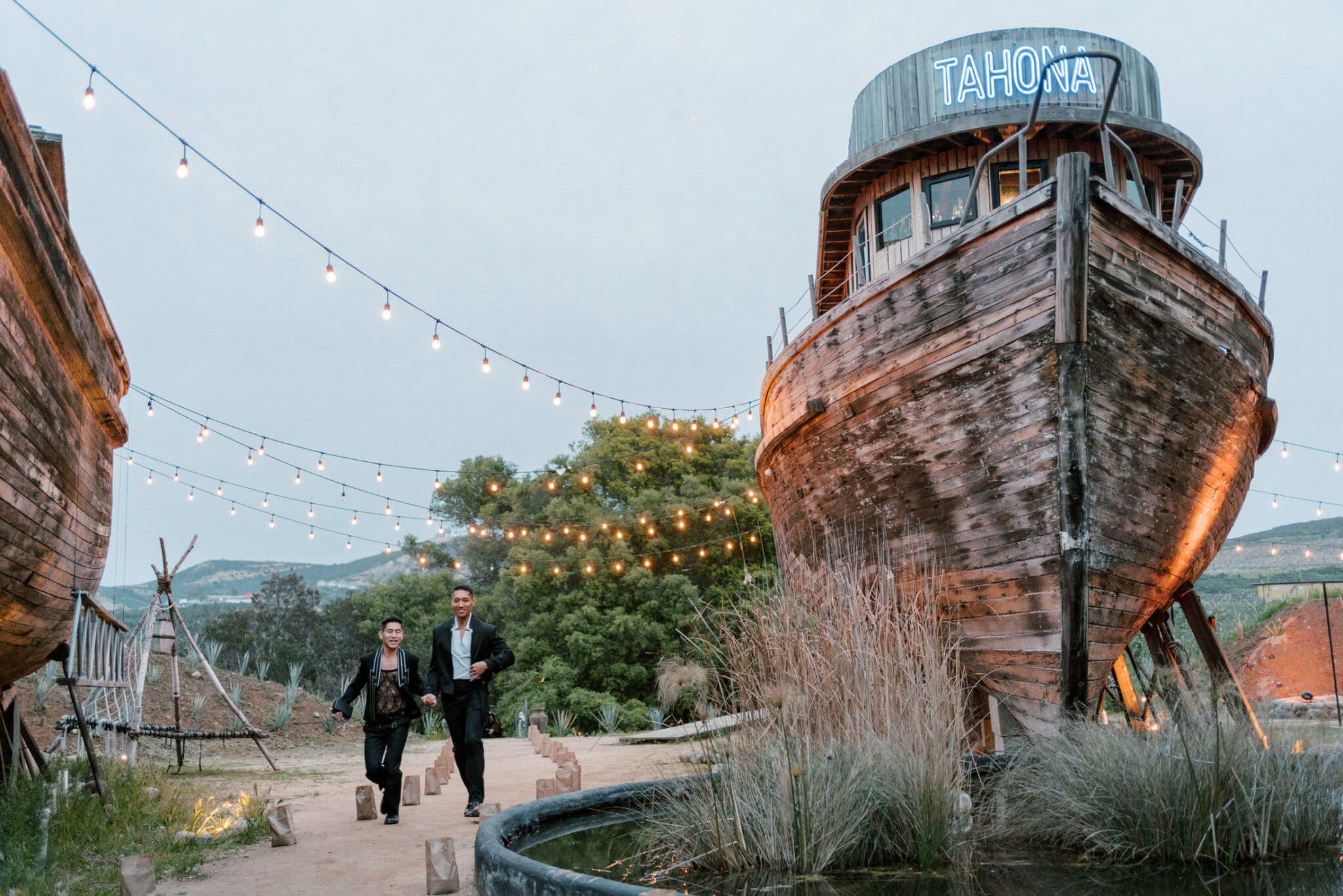 A couple walking hand in hand at dusk through Cuatro Cuatros in Valle de Guadalupe — string lights overhead and the Tahona boat behind them
