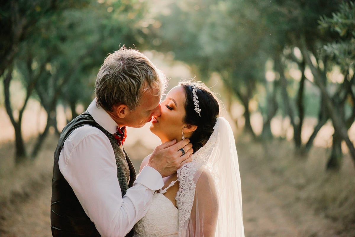 Intimate kiss between bride and groom in olive grove at Corona del Valle — you can feel the lace texture of the dress and the warmth of the light
